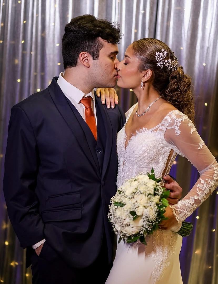 Noivos se beijando diante de cortina prateada com luzes, noiva com buquê de rosas brancas
