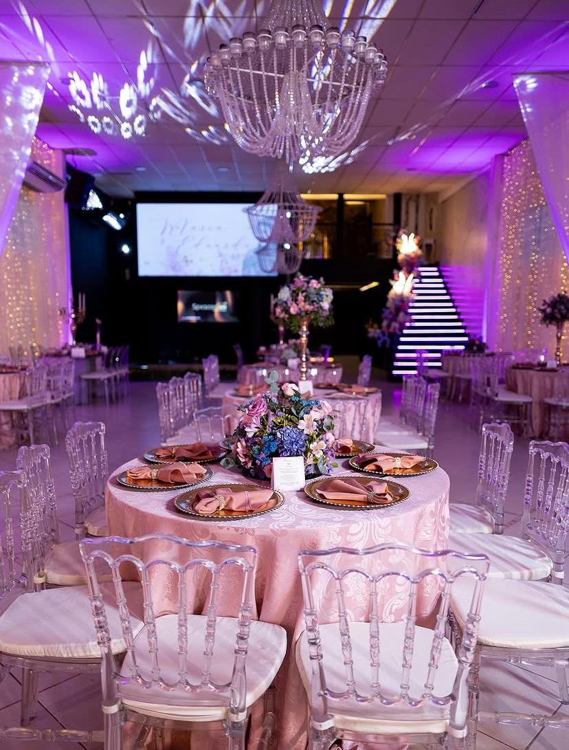 Salão decorado com mesas rosé, cadeiras de cristal, lustres e escadaria iluminada